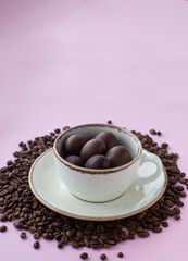 Composition with small chocolate easter eggs in cup surrounded by coffee roasted beans. Easter coffee concept. Mug full of brown eggs on a plate with coffee beans on a pink background with copy space.