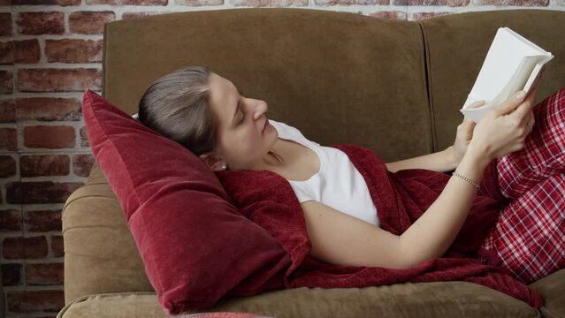 Beautiful young woman lying on sofa in living room and reading a book. People resting at home, hobby, education, female leisure