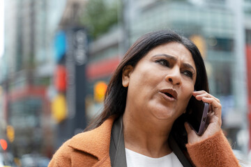 Busy mature Indian woman talking on mobile phone walking on the street. Serious modern female answering call, communication walking on the street