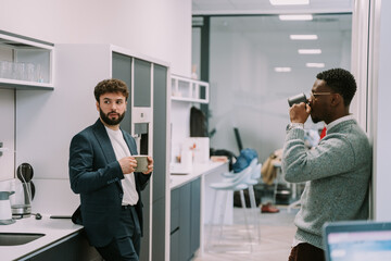 Naklejka na ściany i meble Black male person taking a sip of tea while his white colleague is having a conversation with someone. They are in the office kitchen