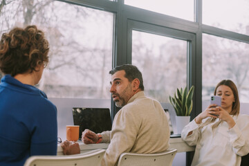 Middle-aged male business person talking to his wife at work while taking a break form work. Their female colleague is checking her phone in the back