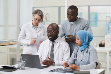 Fototapeta premium African American doctor in white coat presenting online presentation on laptop to colleagues during teamwork in office