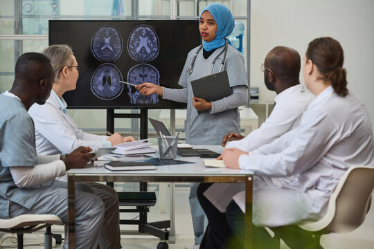 Nurse in hijab pointing at monitor with x-ray images and giving presentation to her colleagues during teamwork in office