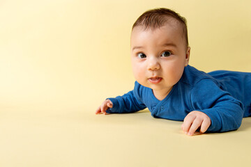 A beautiful little baby in a blue bodysuit, lies on the stomach on a yellow background. Concept, mockup, blank, copy space.