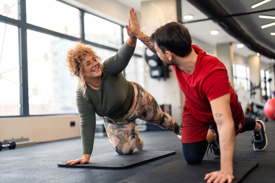 Male personal trainer giving high five support and motivation to smiling young woman fitness client while doing push-ups on exercise mat in gym near panoramic window during individual training.