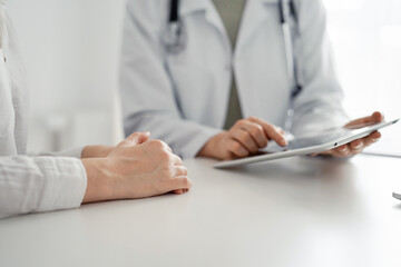 Doctor and patient sitting at the desk in clinic office. The focus is on female physician's hands using tablet computer, close up. Perfect medical service and medicine concept