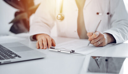 Unknown doctor woman sitting and writing notes at the desk in clinic or hospital office, close up....