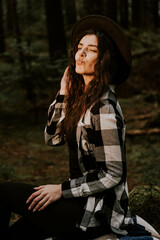 Fototapeta na wymiar Stylish hipster girl in a hat wrapped in a checkered plaid posing in the base of a pine forest.Travel concept