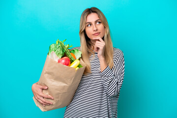 Young Uruguayan woman holding a grocery shopping bag isolated on blue background having doubts and thinking