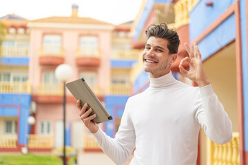 Young caucasian man holding a tablet at outdoors showing ok sign with fingers