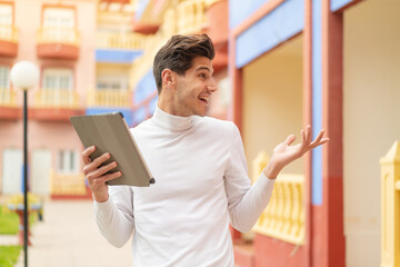 Young caucasian man holding a tablet at outdoors with surprise facial expression