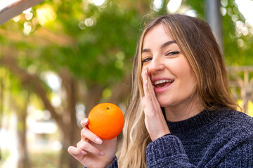 Young pretty Romanian woman holding an orange at outdoors whispering something