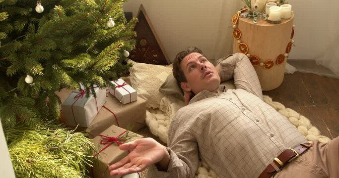 Relaxed american male in casual shirt lying on floor enjoying rest, next to xmas tree. Photo of good mood relaxing male enjoying free time celebrate new year merry christmas at home, thinking