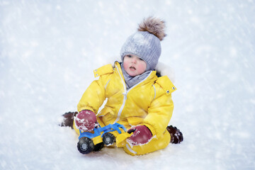 Fototapeta na wymiar Happy toddler baby is playing with a toy in the snow in a yellow snowsuit. Child boy in warm clothes walks in winter park. Kid aged one year six months