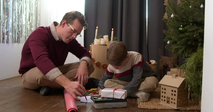 Joyful millennial father drawing painting with adorable little kid son, sitting near decorated Christmas tree, enjoying festive winter holiday New Year celebration time together at home