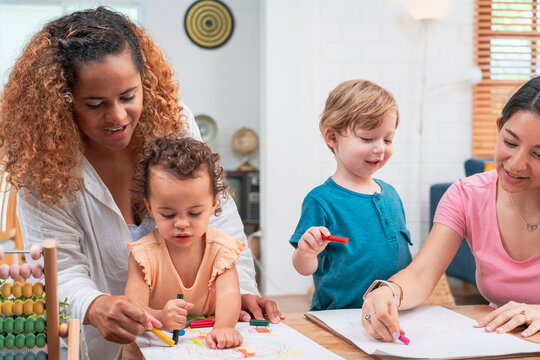 Happy family. Mother and kids together paint. Woman helps the child. Little children with kindergarten teacher drawing at table indoors. Learning and playing