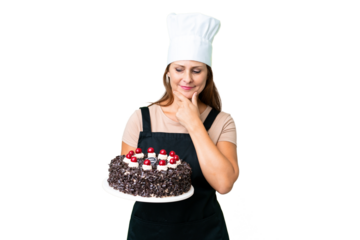 Middle age pastry chef woman holding a big cake over isolated background looking to the side and smiling