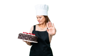 Middle age pastry chef woman holding a big cake over isolated background making stop gesture and disappointed