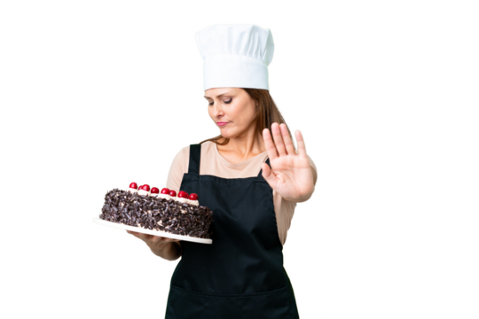 Middle age pastry chef woman holding a big cake over isolated background making stop gesture and disappointed