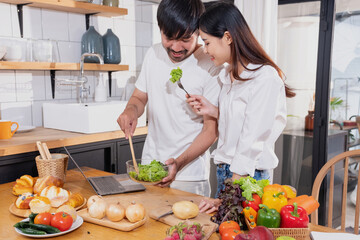Young Asian couple cooking with fruits and vegetables and using laptop in the kitchen To cook food together within the family happily, family concept.