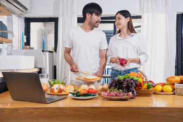 Obraz na płótnie Canvas Young Asian couple cooking with fruits and vegetables and using laptop in the kitchen To cook food together within the family happily, family concept.