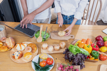 Young Asian couple cooking with fruits and vegetables and using laptop in the kitchen To cook food together within the family happily, family concept.