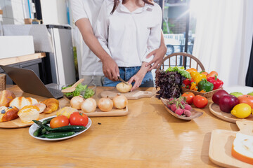 Young Asian couple cooking with fruits and vegetables and using laptop in the kitchen To cook food together within the family happily, family concept.