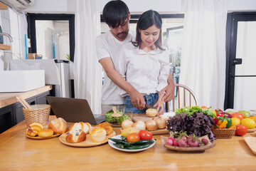 Young Asian couple cooking with fruits and vegetables and using laptop in the kitchen To cook food together within the family happily, family concept.