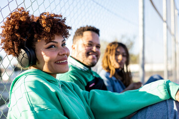 A group of happy and diverse friends gather on a basketball court. The 3 friends are sitting, the girl with afro hair is in the foreground listening to music with headphones. - obrazy, fototapety, plakaty