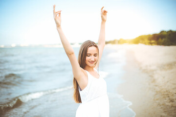 Fototapeta na wymiar Happy smiling woman in free happiness bliss on ocean beach standing with raising hands. Portrait of a multicultural female model in white summer dress enjoying nature