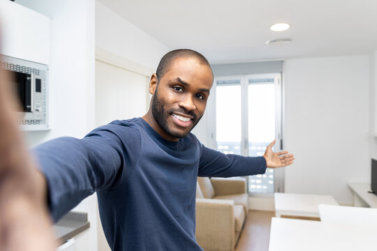 A dark-skinned man takes a selfie in front of a modern smartphone camera with the welcome gesture against the background of a new white apartment he rented, celebrating the last mortgage payment.