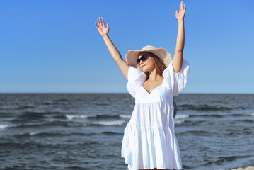 Fototapeta na wymiar Happy blonde woman is on the ocean beach in a white dress and sunglasses, raising hands