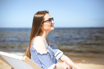 Fototapeta na wymiar Happy young brunette woman relaxing on a wooden deck chair at the ocean beach while smiling, and wearing fashion sunglasses. The enjoying vacation concept