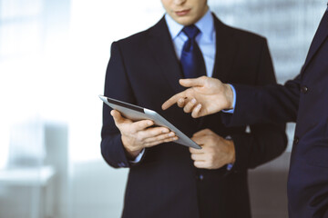 Business people use a tablet computer for discussion of their new project, standing in a modern office. Unknown businessman or male entrepreneur with a colleague at workplace.