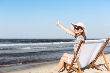 Happy brunette woman sitting on a wooden deck chair at the ocean beach while showing Thumb up