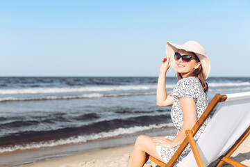 Obraz na płótnie Canvas Happy brunette woman wearing sunglasses while relaxing on a wooden deck chair at the ocean beach
