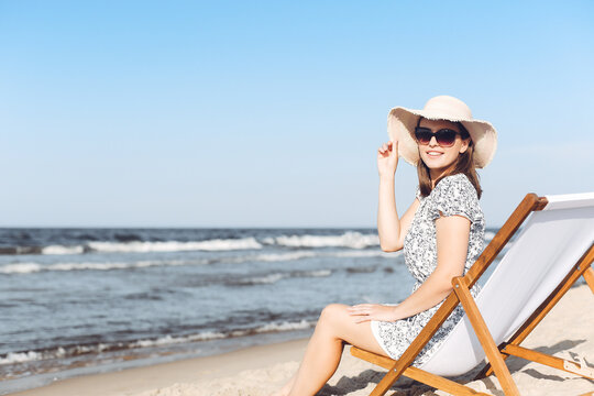 Happy brunette woman wearing sunglasses while relaxing on a wooden deck chair at the ocean beach