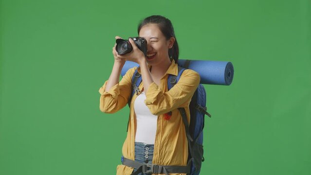 Asian Female Hiker With Mountaineering Backpack Using A Camera Taking Picture While Standing On Green Screen Background In The Studio
