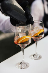 Bartender adding ice cubes from into Aperol spritz cocktail glass. Barman serving orange liqueur cocktail in a wine glass on a bar counter and slice of orange.