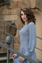 A beautiful, young girl with dark hair in a gray dress stands on the carved stairs of the palace. Grace and elegance.setting sun