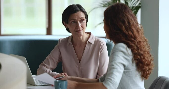 Mature business mentor woman talking to younger female employee, intern at workplace, speaking, teaching, explaining work tasks, asking questions, smiling, laughing, sitting at table with laptop