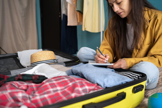 Latin woman making check list of things to pack for travel, writing notes and packing suitcase to vacation sitting on the bed, prepare clothes into luggage. Travel vacation