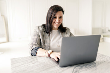 An online teacher uses a laptop of a brunette woman in stylish glasses and a jacket indoors, smiling, showing her teeth, happy with every working day.