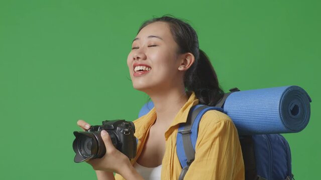Close Up Side View Of Asian Female Hiker With Mountaineering Backpack Smiling And Holding A Camera In Her Hands Then Looking Around While Standing On Green Screen Background In The Studio
