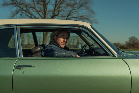 Man With Blond Hair In Jeans Jacket Sits On Passenger Side Of A Vintage American Muscle Car Looking Out The Window.