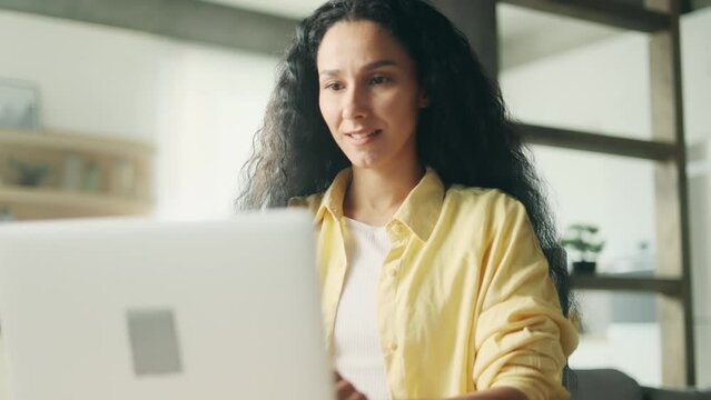 Smiling young hispanic woman relaxing at home or working online typing browsing products in internet store reading news checking email or social media profile on laptop and looking at camera indoors