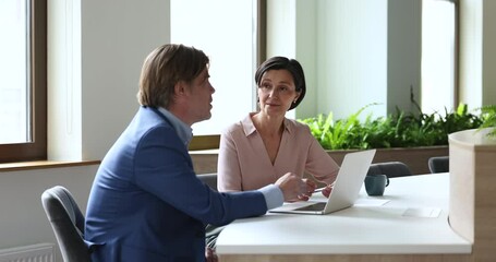 Mature couple of coworkers talking at workplace with laptop. Businesswoman negotiating with deal partner, colleague, employee, sitting at table, telling work tasks, speaking - Powered by Adobe