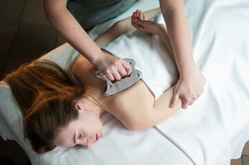 Hands of the masseur are close-up using a metal massage tool to massage the neck of a woman lying on a couch. Health concept, body care, skin care, wellness