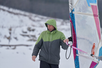 A middle-aged man, a snowsurfer, stands on the ice of a frozen lake and waits for a woman. Preparing to ride a sailboard in the snow on a cloudy winter day.