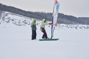 A middle-aged woman, a snowsurfer, starts moving on a sailboard, while a man watches. A middle-aged man and woman go snowsurfing on a cloudy winter day.